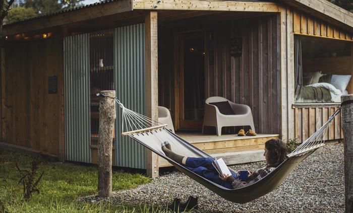 A woman lies in a hammock whilst reading a book. In the background is a wooden cabin with some lounge chairs on a deck and a large window with an unma